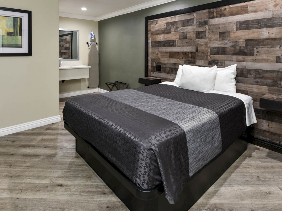 A modern bedroom with a wooden accent wall, a bed with gray bedding, a nightstand, a framed picture, and a sink area in the background.