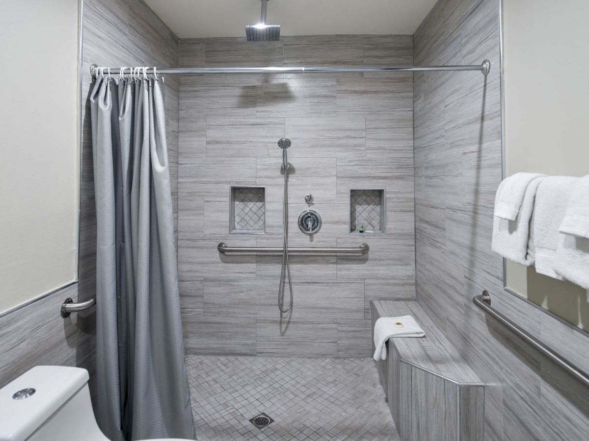 A bathroom with a walk-in shower, gray curtain, shower bench, two niches, rainfall shower head, grab bars, and a toilet. Towels are on the right side.