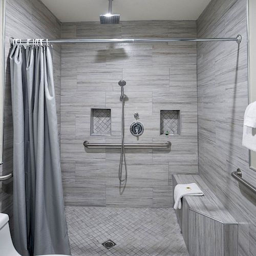 A modern bathroom with a walk-in shower, grey tiles, a showerhead, a bench, towel railings with white towels, and a toilet on the left.
