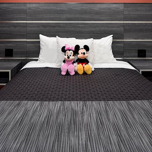 A neatly made bed with gray and black bedding, featuring two stuffed toys (Mickey and Minnie Mouse) propped against white pillows on it.