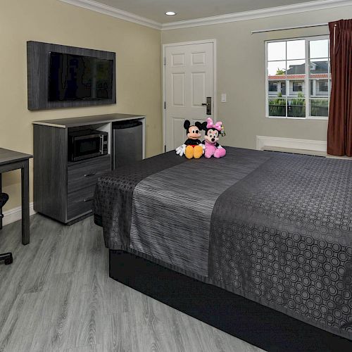 A well-kept room with a bed, flat-screen TV, desk, chair, and a figurine of Mickey Mouse and Minnie Mouse on the bed.