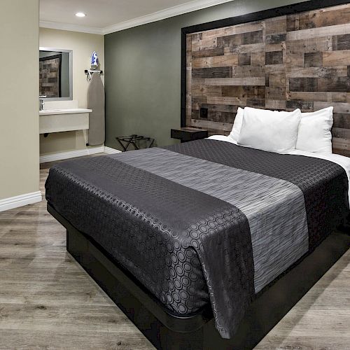 A modern bedroom featuring a bed with dark bedding, a rustic wooden headboard, artwork, and an adjacent sink area with a mirror.