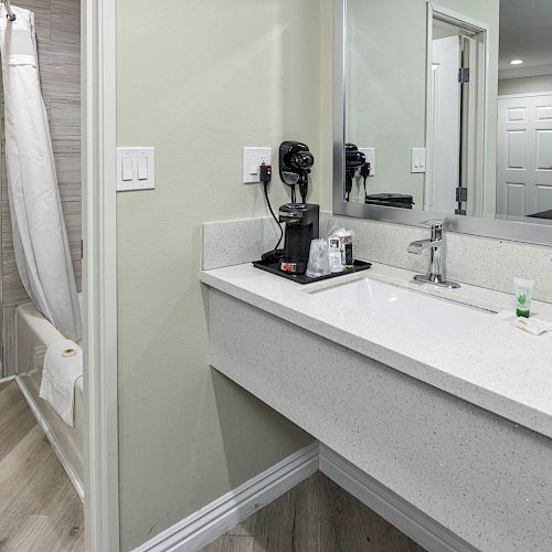 The image shows a modern bathroom with a toilet, a vanity sink with a mirror, and a shower with a white curtain. A hairdryer and toiletries are on the counter.