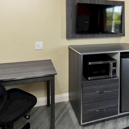 The image shows a desk with a black chair next to a cabinet holding a microwave, a small refrigerator, and a mounted TV on the wall.
