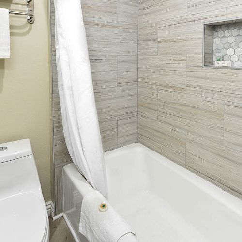 A modern bathroom with a white toilet, a bathtub with a white curtain, and a beige tiled wall with a built-in shelf.
