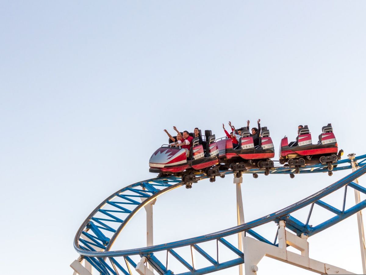 A red roller coaster car full of people with hands raised is navigating a blue and white track against a clear sky.