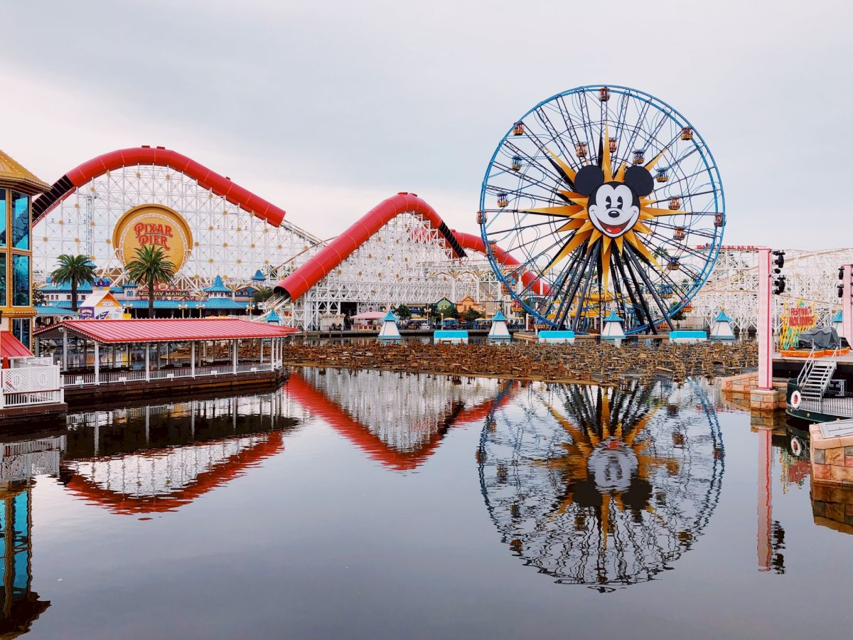 The image shows an amusement park with a prominent Ferris wheel featuring a cartoon face and a roller coaster, both reflected in a calm body of water.