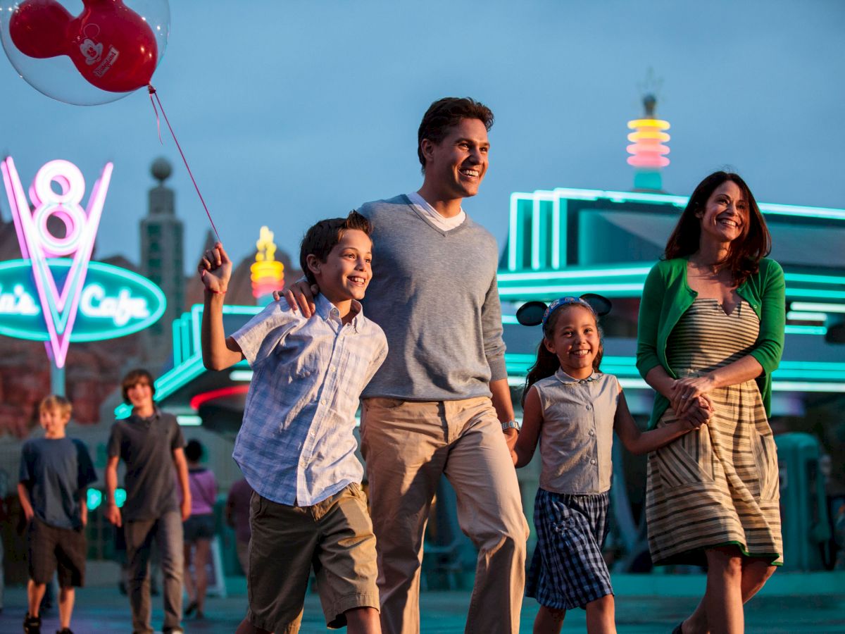 A family of four enjoys a night out, walking in front of a bright, neon-lit diner, with the son holding red balloons, smiling together.