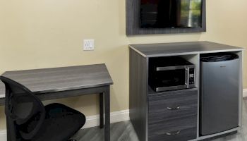 The image shows a small office area with a desk, chair, mounted TV, microwave, and a mini-fridge in a corner of a room.