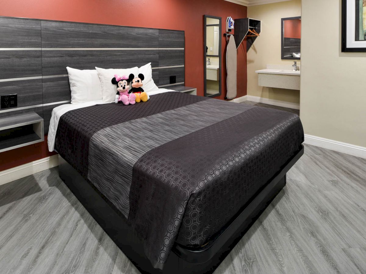 A bedroom with a bed adorned with gray and black bedding, stuffed toys on the pillows, a bathroom sink area, and a wall-mounted TV.