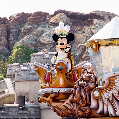 The image shows a Mickey Mouse character wearing a crown on a parade float with scenic rocky hills in the background.