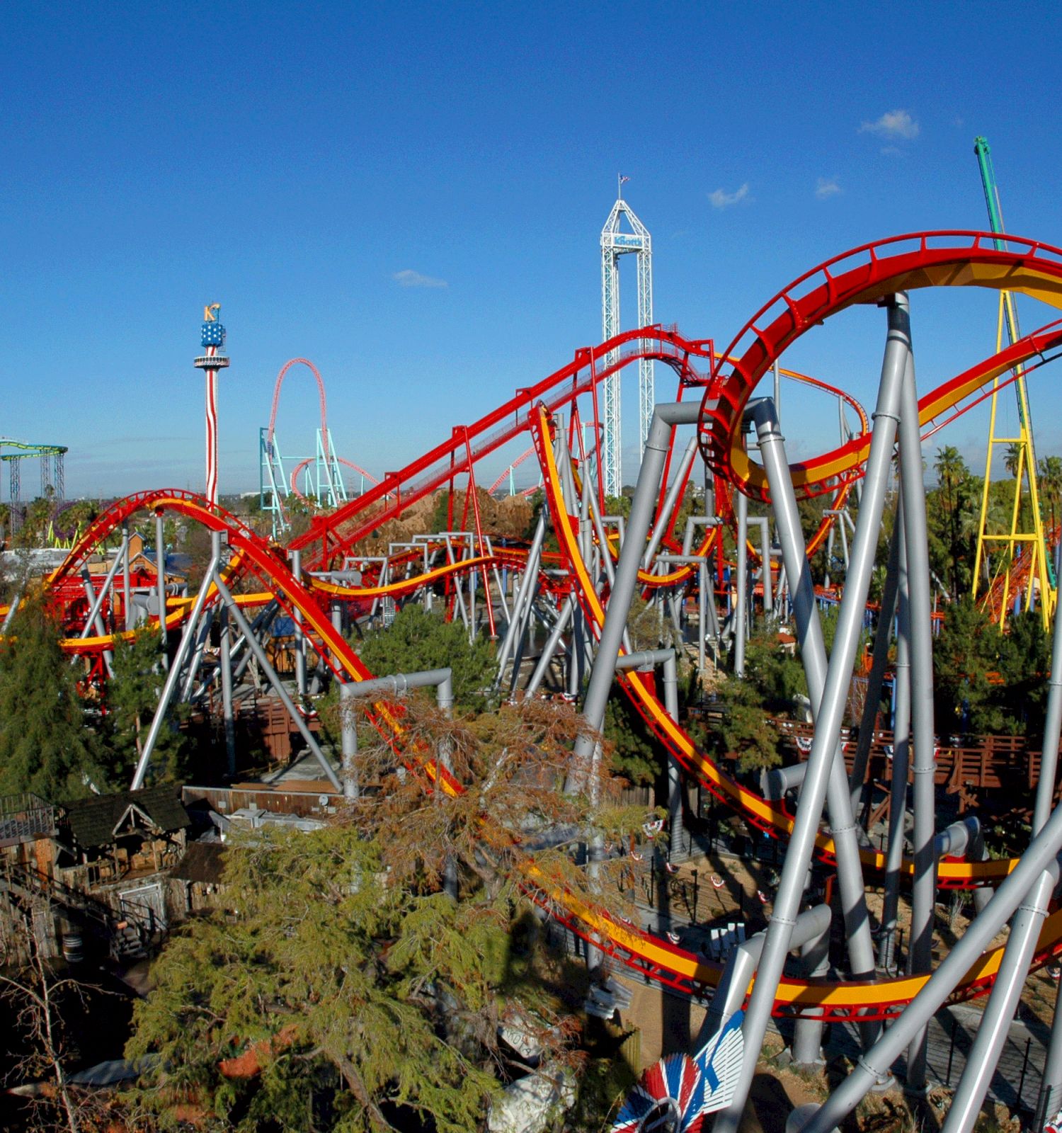 The image shows a vibrant amusement park with several roller coasters, including a prominent red and yellow one, surrounded by greenery and other attractions.