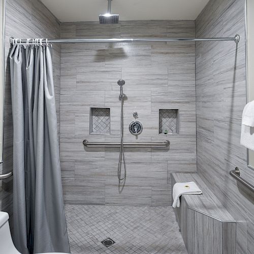 A modern, accessible shower with gray tiles, a shower curtain, grab bars, a shower bench, and a toilet nearby. Towels are hung on a rack.